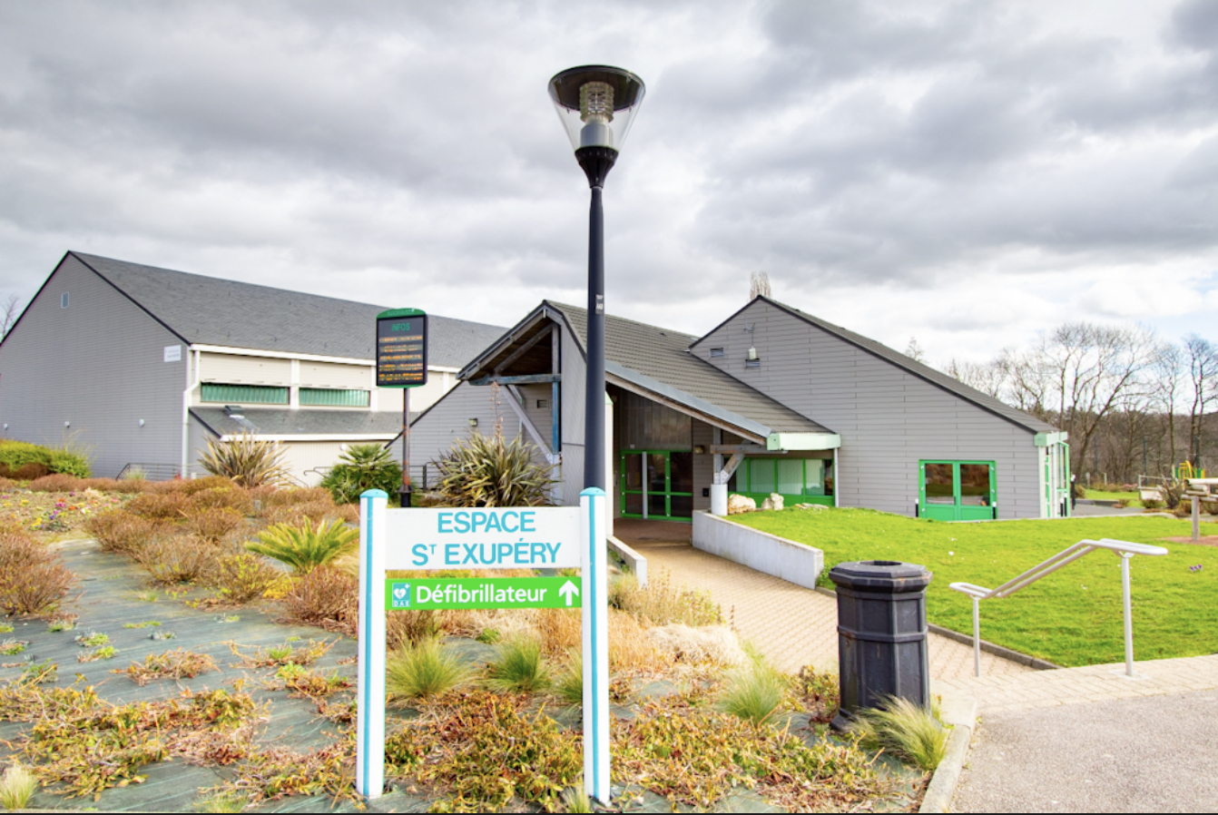 Vue de la salle Saint Exupéry de la commune avec le panneau de signalisation et les bâtiments en arrière plan